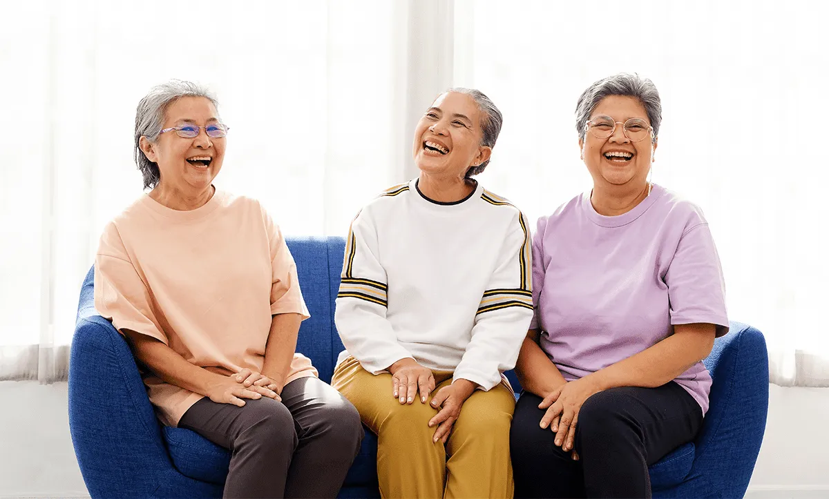 Three older women with short gray hair sit together on a blue sofa, smiling and laughing, in front of a bright window; they wear colorful clothes and embody the joyful, relaxed spirit of Retirement Living at MorningStar Senior Living Kirkland.