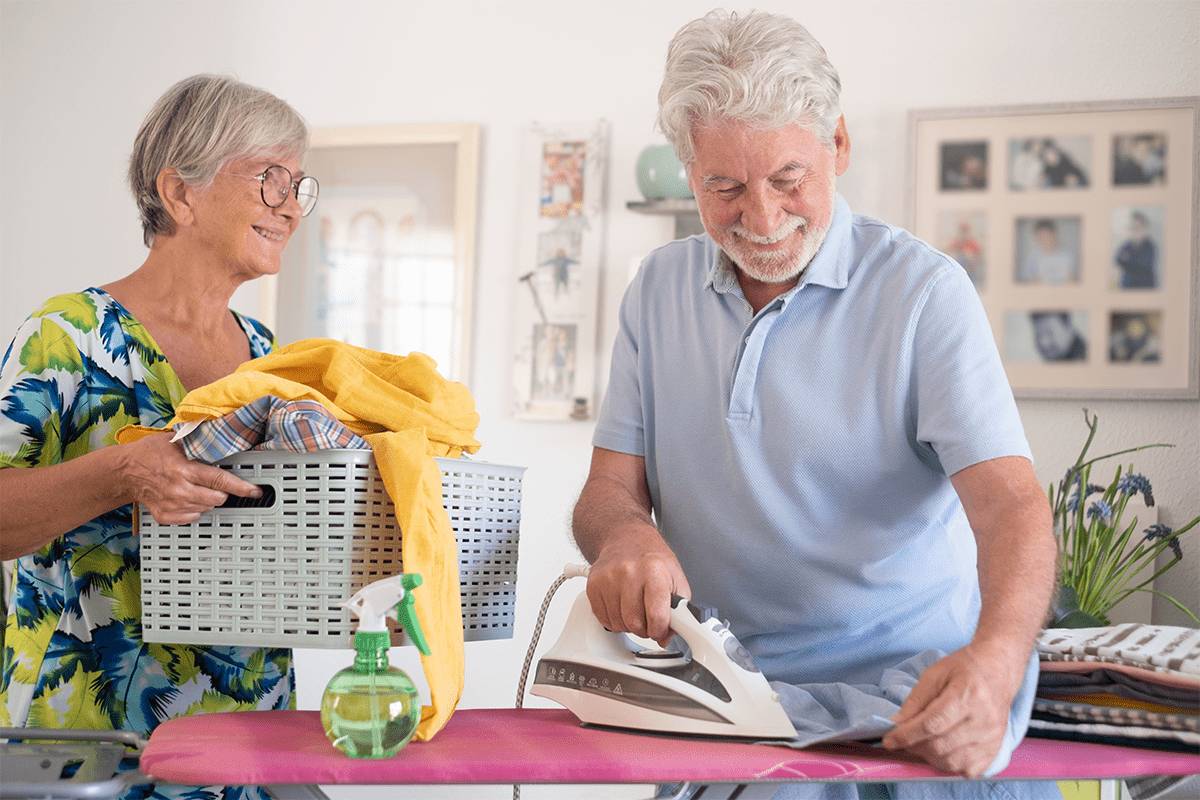An older woman holds a laundry basket and smiles at an older man ironing clothes in a bright, cozy room at MorningStar Senior Living Kirkland. They appear happy, doing housework together and enjoying retirement living.