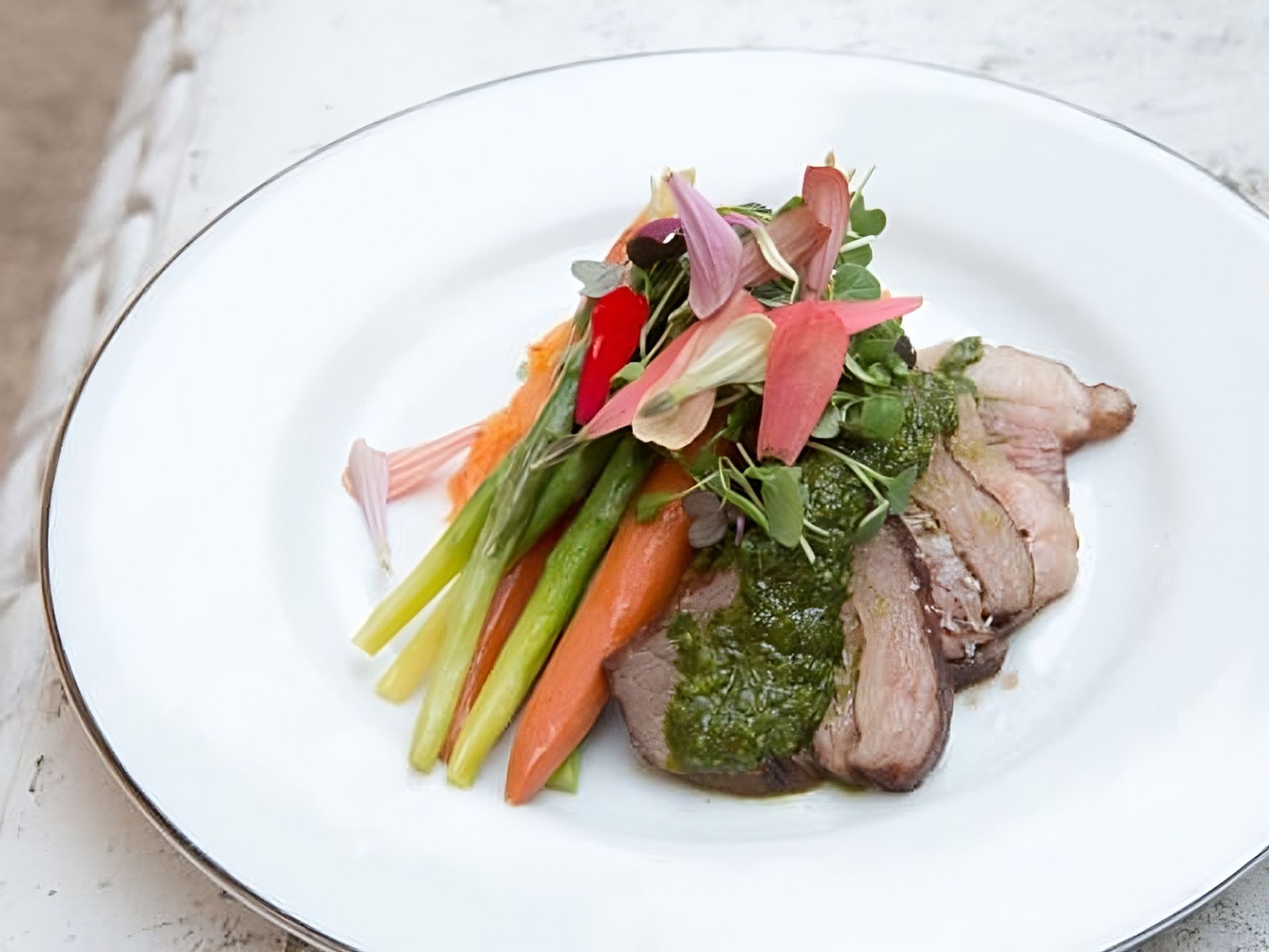 A plated dish at MorningStar Senior Living Kirkland features slices of cooked meat topped with green sauce, carrots, asparagus, and microgreens, garnished with edible flower petals—a vibrant option for refined retirement living.