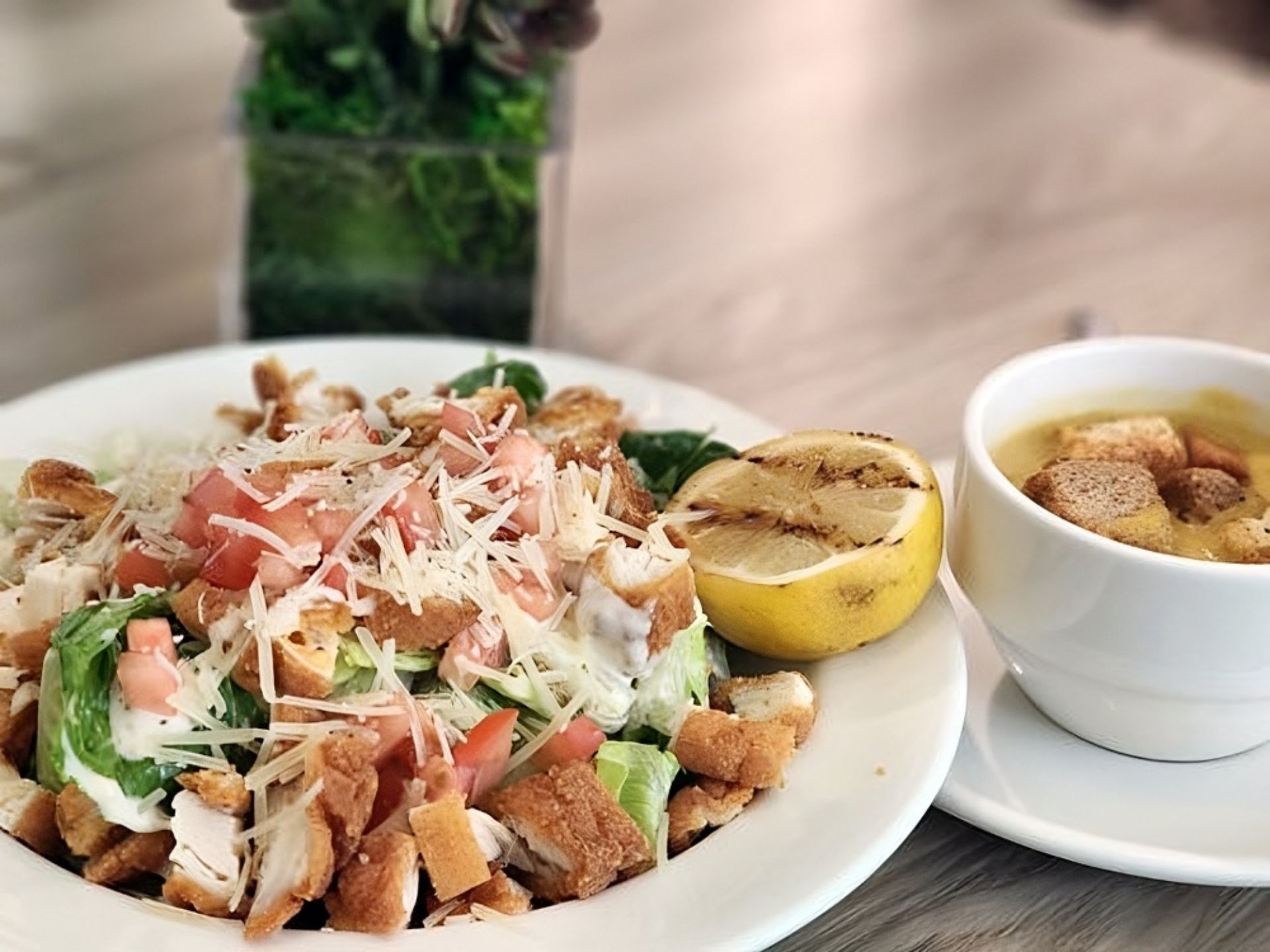 A plate of Caesar salad with croutons, diced tomatoes, grilled chicken, and a grilled lemon sits beside a cup of soup. A small plant decorates the background—just one example of the delicious meals at MorningStar Senior Living Kirkland.