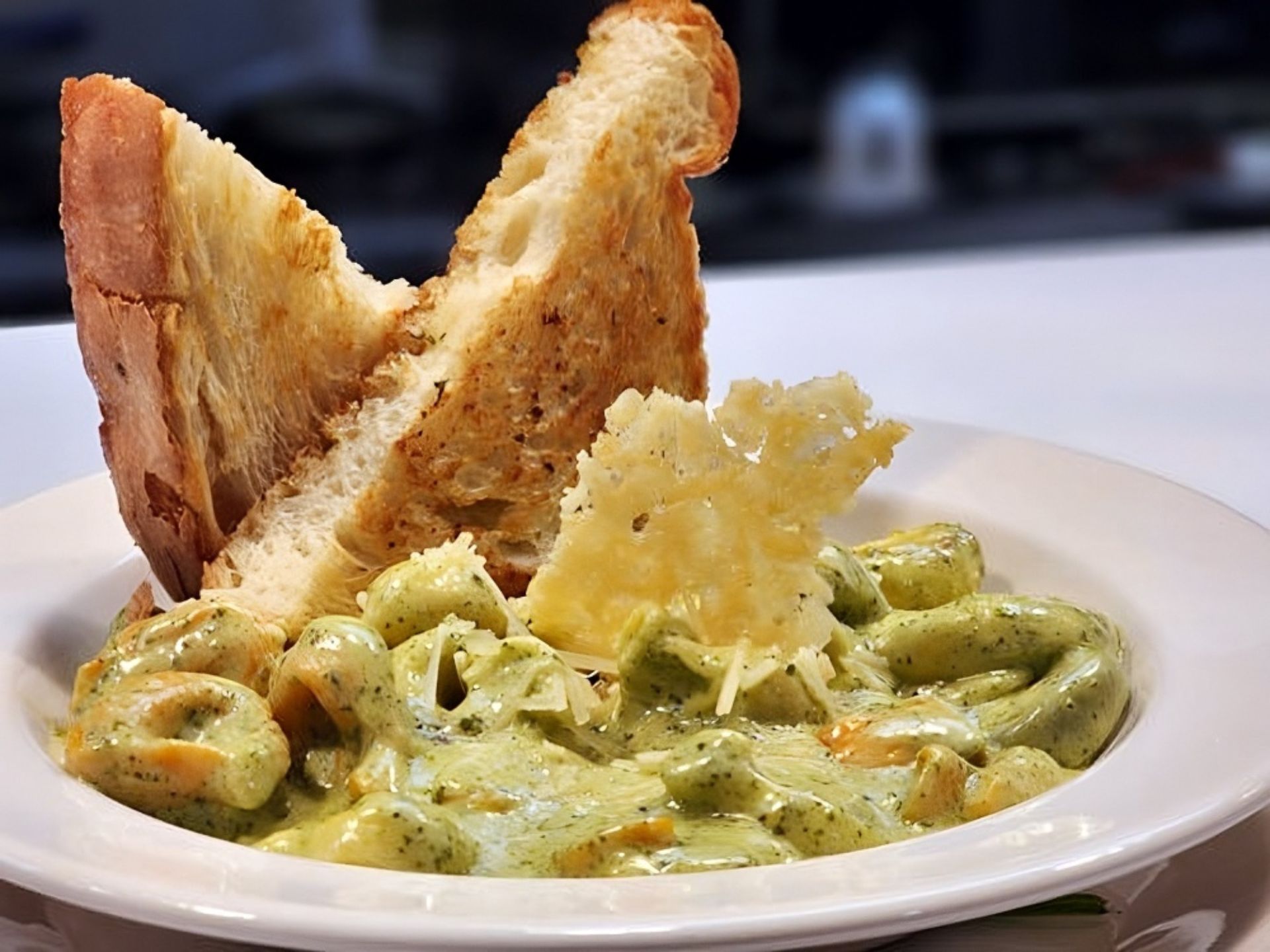 At MorningStar Senior Living Kirkland, a white bowl filled with creamy green pesto pasta, topped with shredded cheese and a crispy cheese garnish, is served alongside two slices of toasted bread standing upright in the bowl—perfect for retirement living.