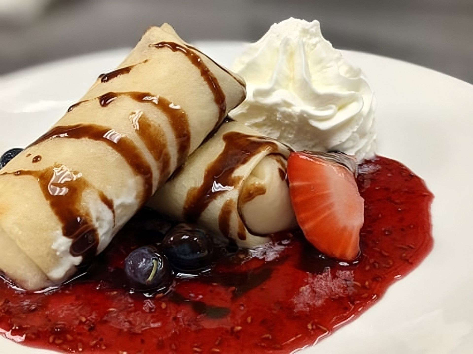 At MorningStar Senior Living Kirkland, enjoy two crepes drizzled with chocolate sauce, served with whipped cream, fresh blueberries, a strawberry slice, and raspberry sauce on a white plate—a delicious touch to retirement living.