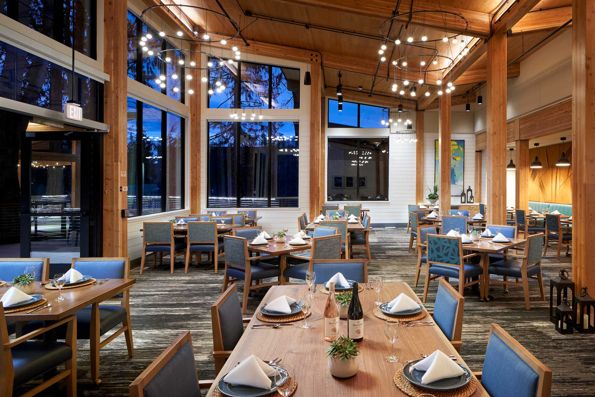 A modern, spacious restaurant at MorningStar Senior Living Kirkland features wooden beams, large windows, and blue chairs. Tables are set with plates, napkins, wine glasses, and small plants under warm lighting—ideal for refined retirement living.