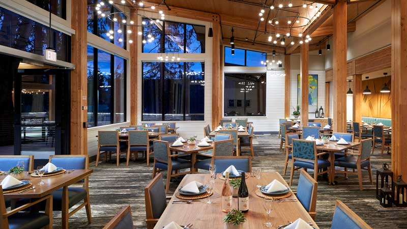 Modern restaurant interior at MorningStar Senior Living Kirkland, featuring wooden beams, large windows revealing the evening sky, blue cushioned chairs, neatly set tables with wine glasses, and string lights for a warm retirement living ambiance.