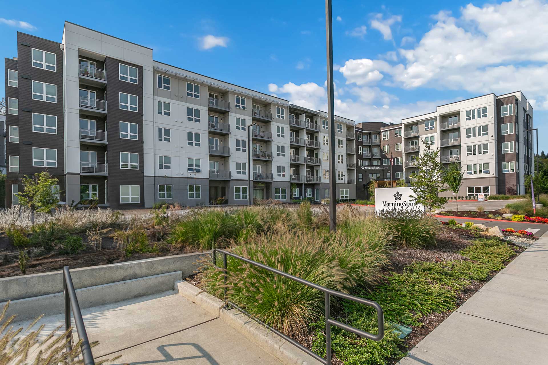Modern four-story apartment building with balconies, surrounded by landscaped greenery and a sidewalk. A sign near the entrance displays MorningStar Senior Living Kirkland, offering an inviting retreat for retirement living under blue, scattered-cloud skies.