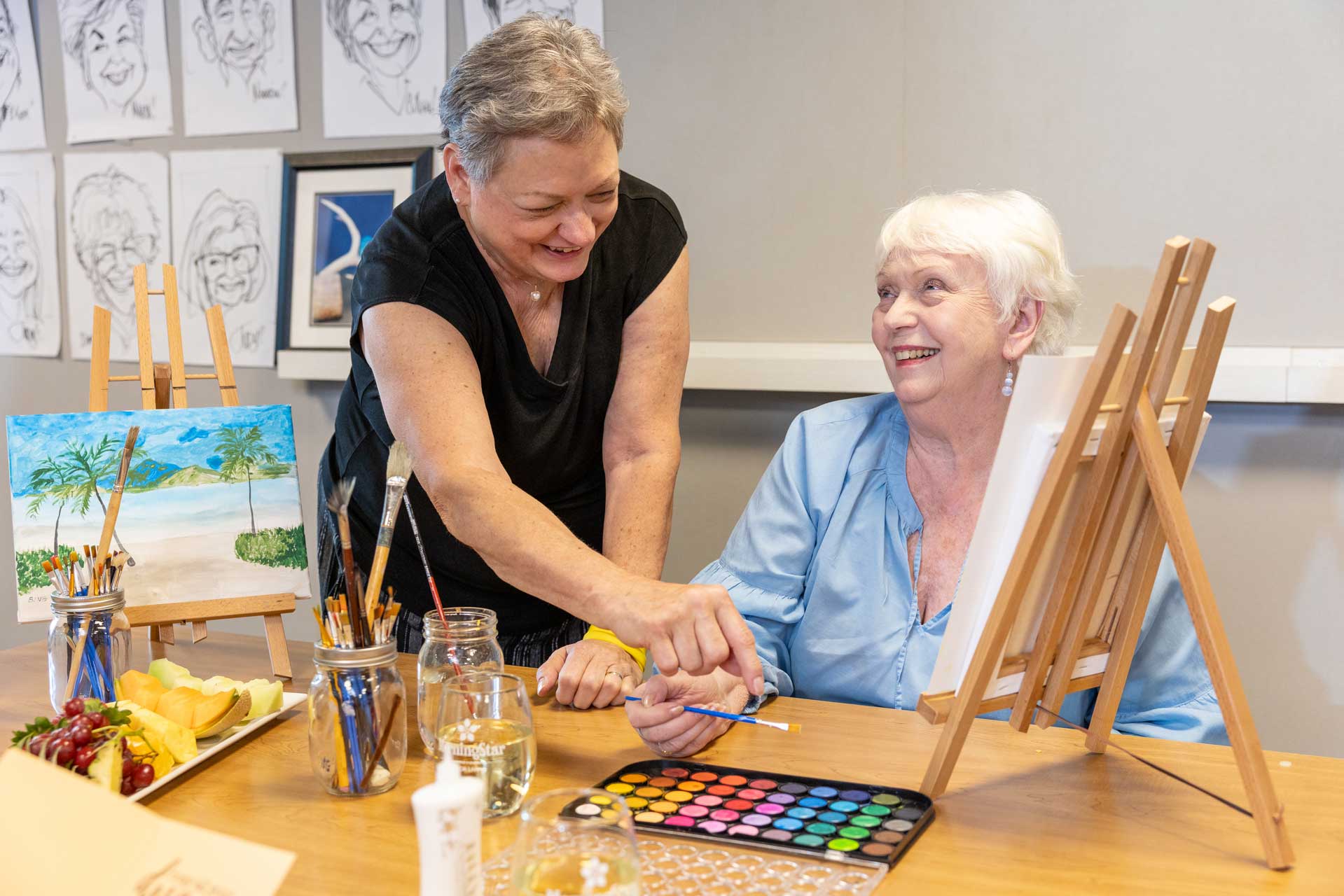Two older women smile while painting together at a table with art supplies at MorningStar Senior Living Kirkland. One helps the other with a paintbrush, surrounded by sketches and a finished beach landscape—a joyful moment in retirement living.