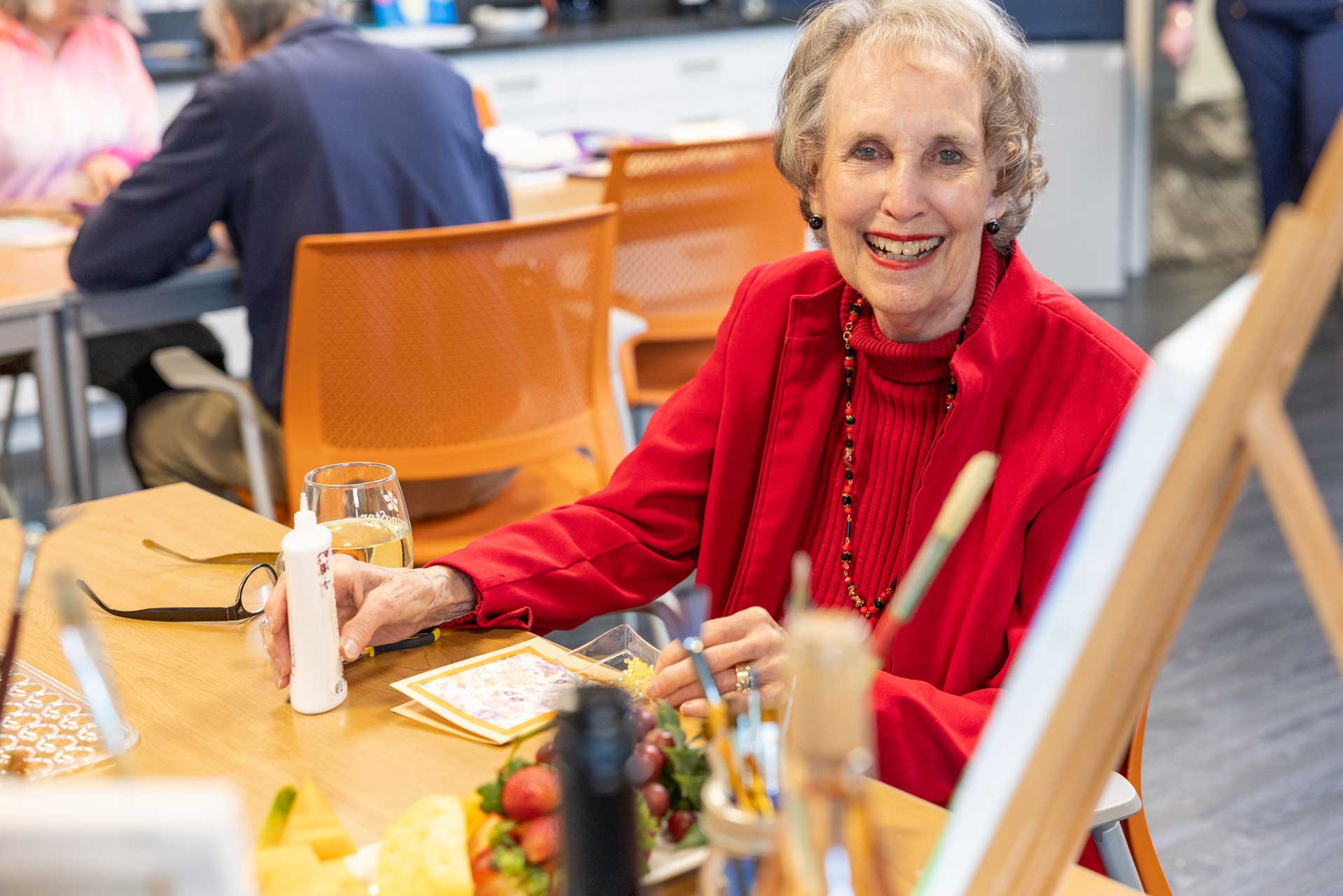 An elderly woman in a red jacket smiles while sitting at a table set with art supplies, fruit, and water at MorningStar Senior Living Kirkland, suggesting a vibrant group activity or gathering in retirement living.