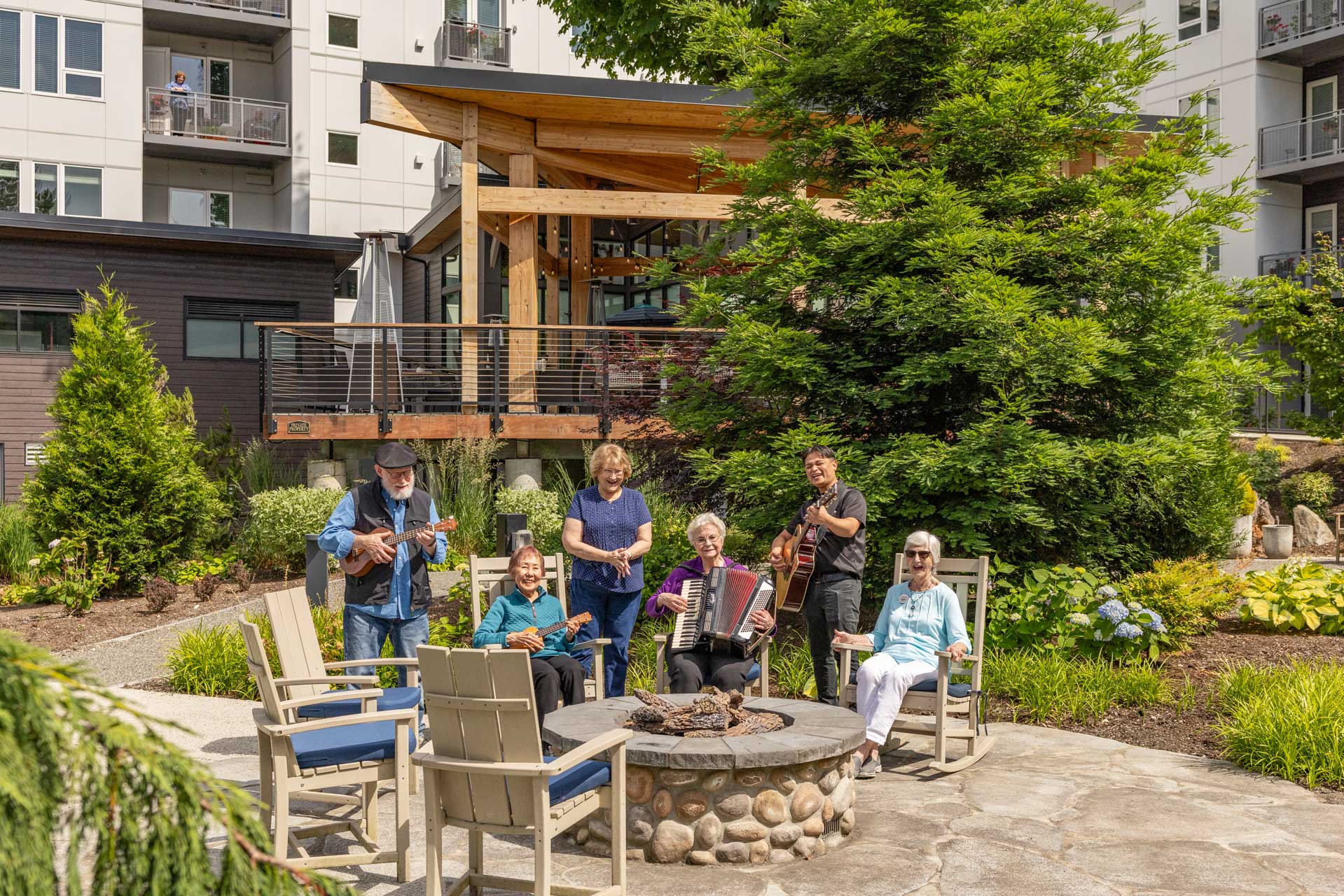 A group of six seniors, some playing musical instruments, sit and stand around a stone fire pit in a landscaped courtyard at MorningStar Senior Living Kirkland, enjoying vibrant retirement living outside a modern apartment building on a sunny day.
