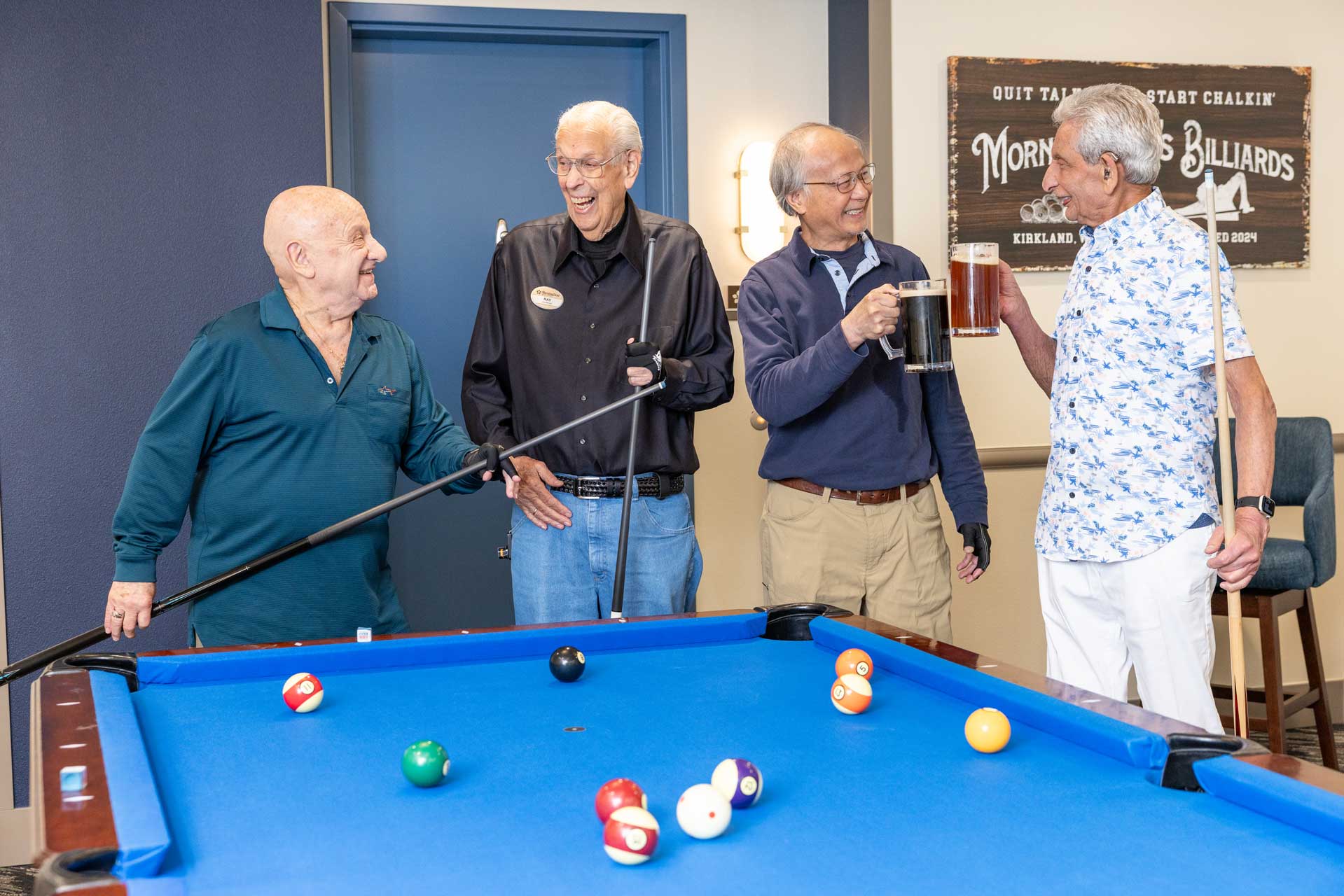 Four older men stand around a blue pool table at MorningStar Senior Living Kirkland, smiling and laughing. One holds a cue stick, another has a large mug of beer. A billiards sign hangs on the wall behind them; pool balls are scattered across the table—retirement living at its best.