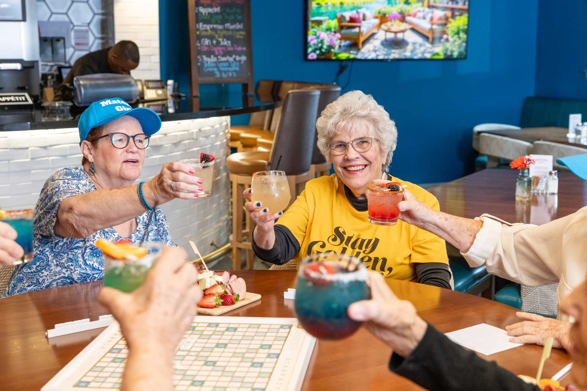 A group of elderly women at MorningStar Senior Living Kirkland sit around a table, smiling and raising colorful drinks in a toast. A Scrabble board and snacks are on the table in this cheerful retirement living lounge.