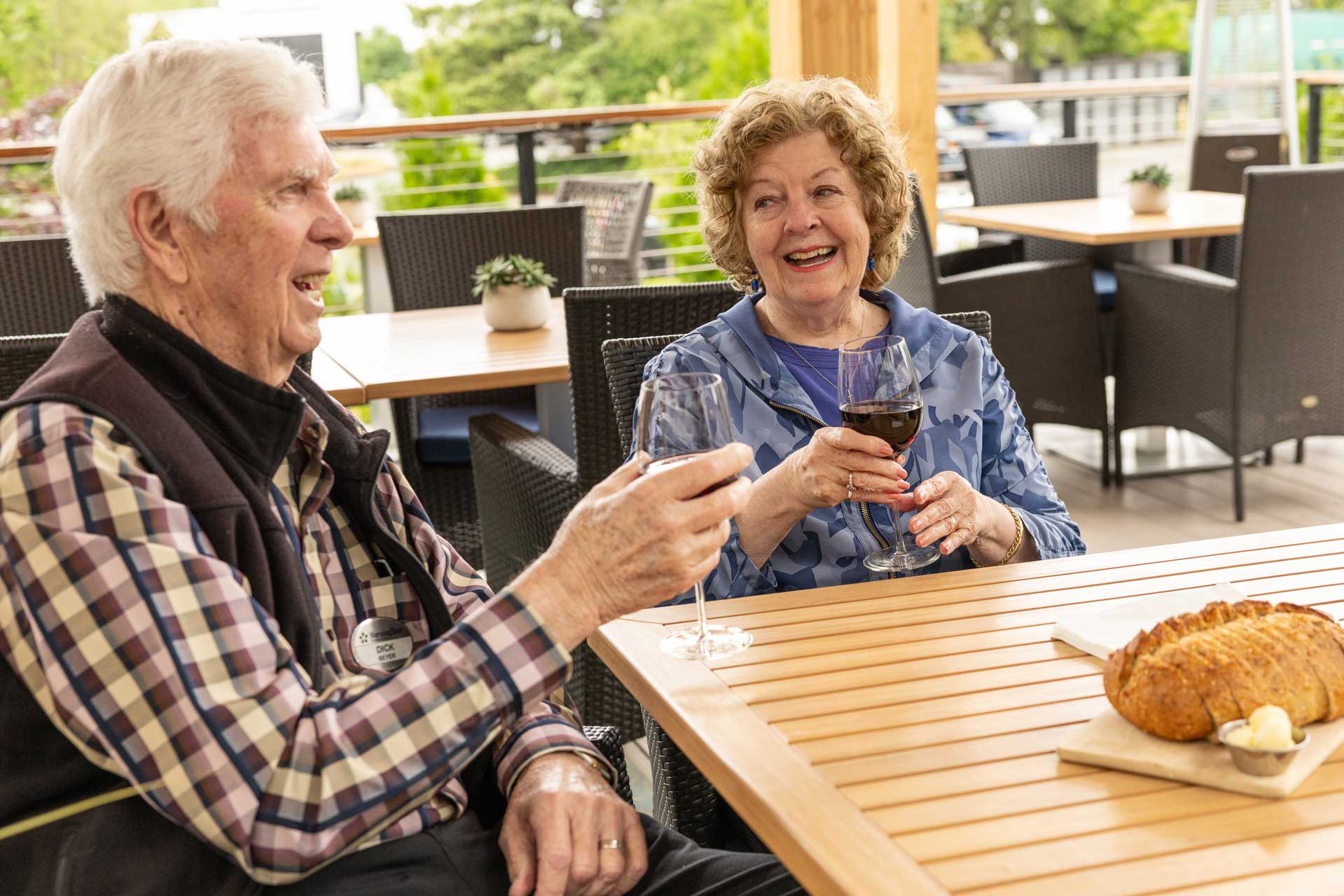 Two older adults sit at an outdoor table at MorningStar Senior Living Kirkland, smiling and holding glasses of red wine. A loaf of bread with butter is between them, enjoying relaxed retirement living with greenery in the background.