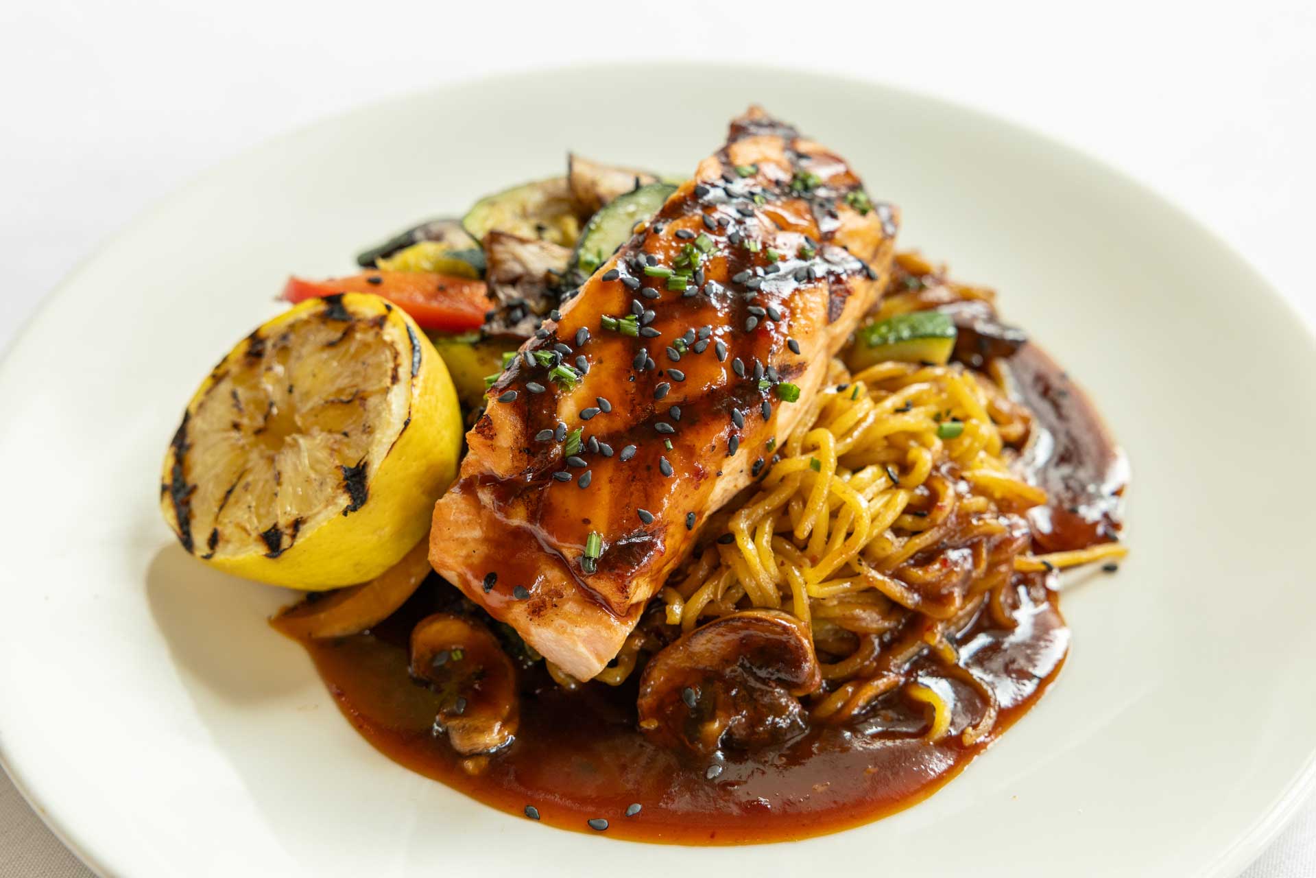 Grilled salmon fillet with black sesame seeds on top, served over noodles and vegetables in a rich brown sauce, accompanied by a grilled lemon half—a delicious option offered at MorningStar Senior Living Kirkland for elevated retirement living.