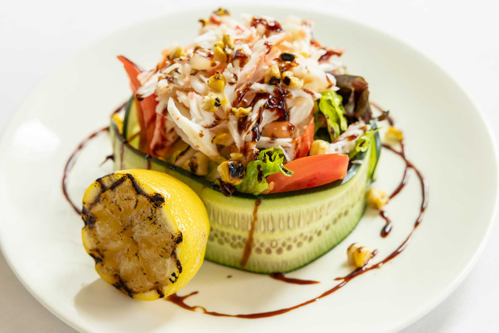 A fresh salad with shredded crab, lettuce, and tomato wrapped in cucumber slices, garnished with chopped nuts and balsamic glaze, served with charred lemon—an elegant dish perfect for Retirement Living at MorningStar Senior Living Kirkland.