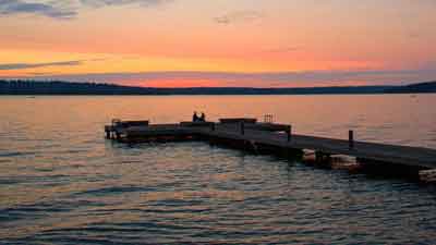 A wooden pier extends over calm water at sunset near MorningStar Senior Living Kirkland, with two people sitting at the end, silhouetted against a colorful sky—a serene scene perfect for retirement living.