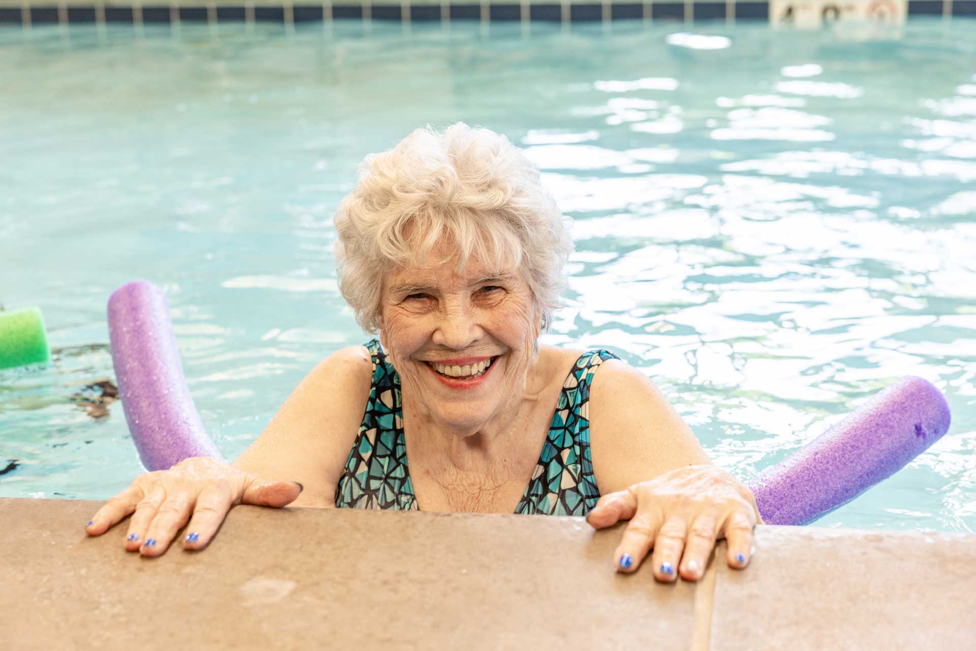 An older woman with short white hair smiles while holding onto the edge of a swimming pool at MorningStar Senior Living Kirkland. She wears a patterned swimsuit, colorful pool noodles surround her, and her nails are painted blue—embracing vibrant retirement living.