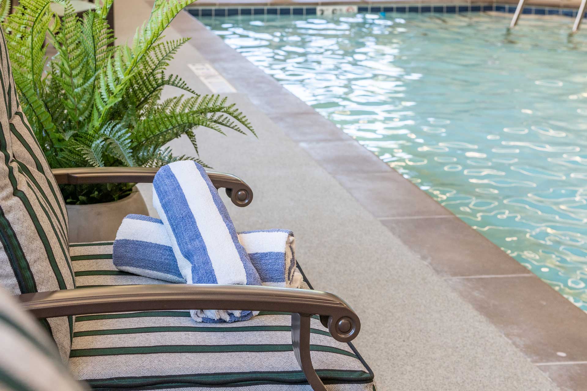 A striped cushioned chair by an indoor pool at MorningStar Senior Living Kirkland holds two rolled blue and white towels, with a potted fern nearby and the pool's water and tile edge visible in the background.
