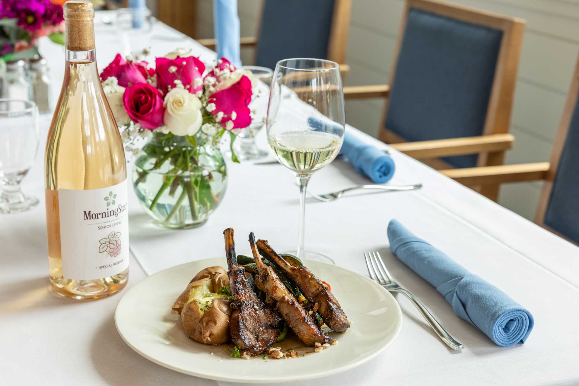 A plate with grilled lamb chops, baked potato, and garnish sits on a white tablecloth alongside a glass and bottle of white wine at MorningStar Senior Living Kirkland, offering elegant retirement living in a refined dining setting.