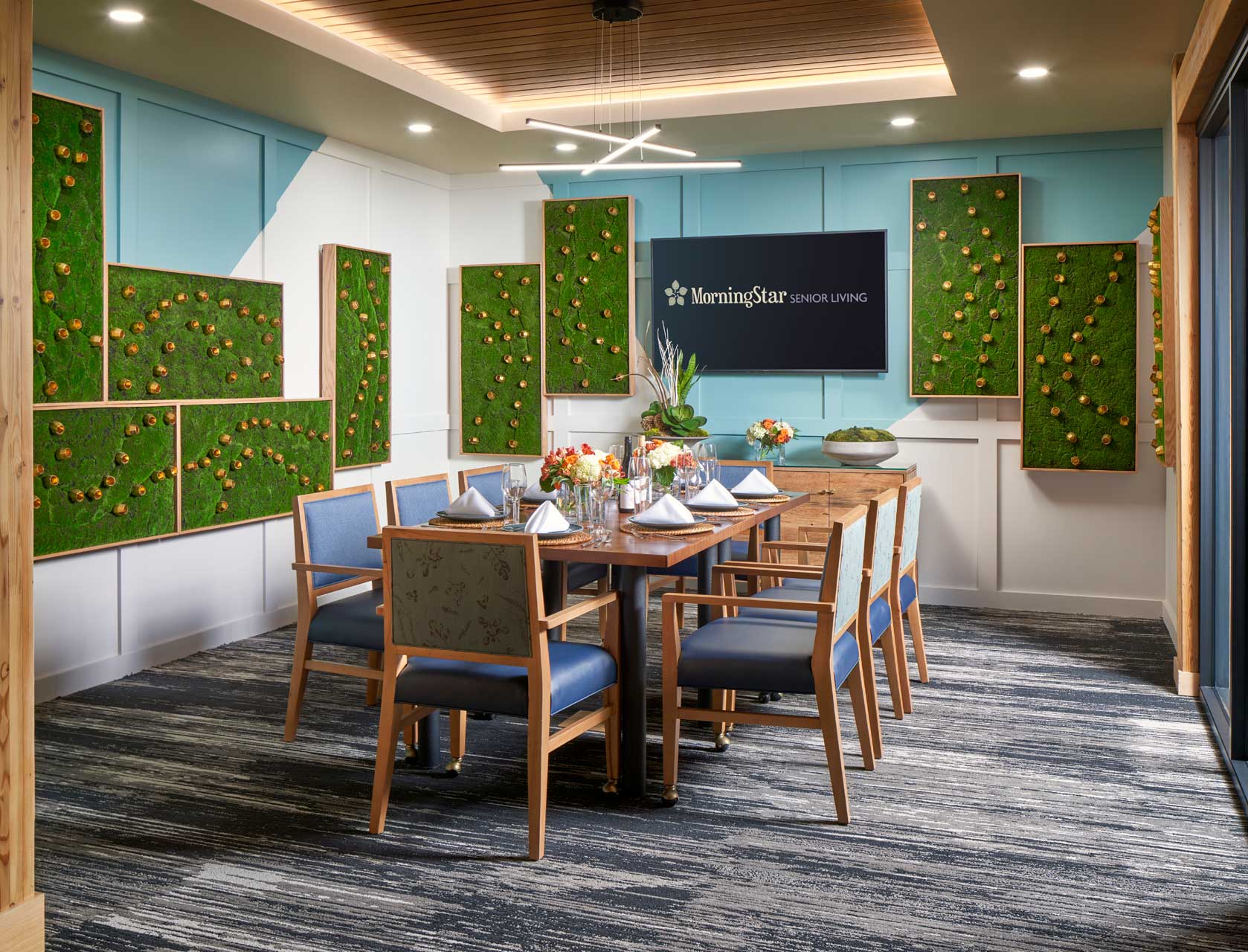 A modern dining room at MorningStar Senior Living Kirkland features a wooden table set for eight, blue cushioned chairs, floral centerpieces, a TV displaying 