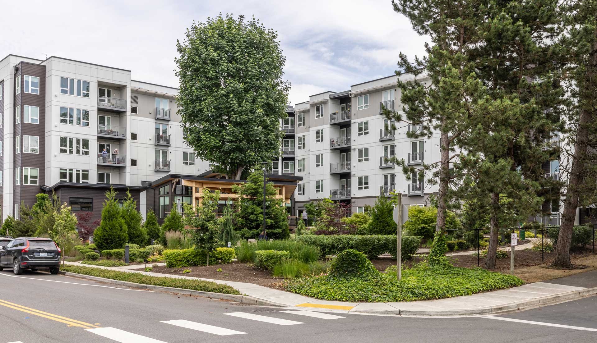 Modern apartment building with balconies, surrounded by lush green landscaping, trees, and gardens; a parked car and street with a crosswalk are in the foreground—ideal for comfortable Retirement Living at MorningStar Senior Living Kirkland.