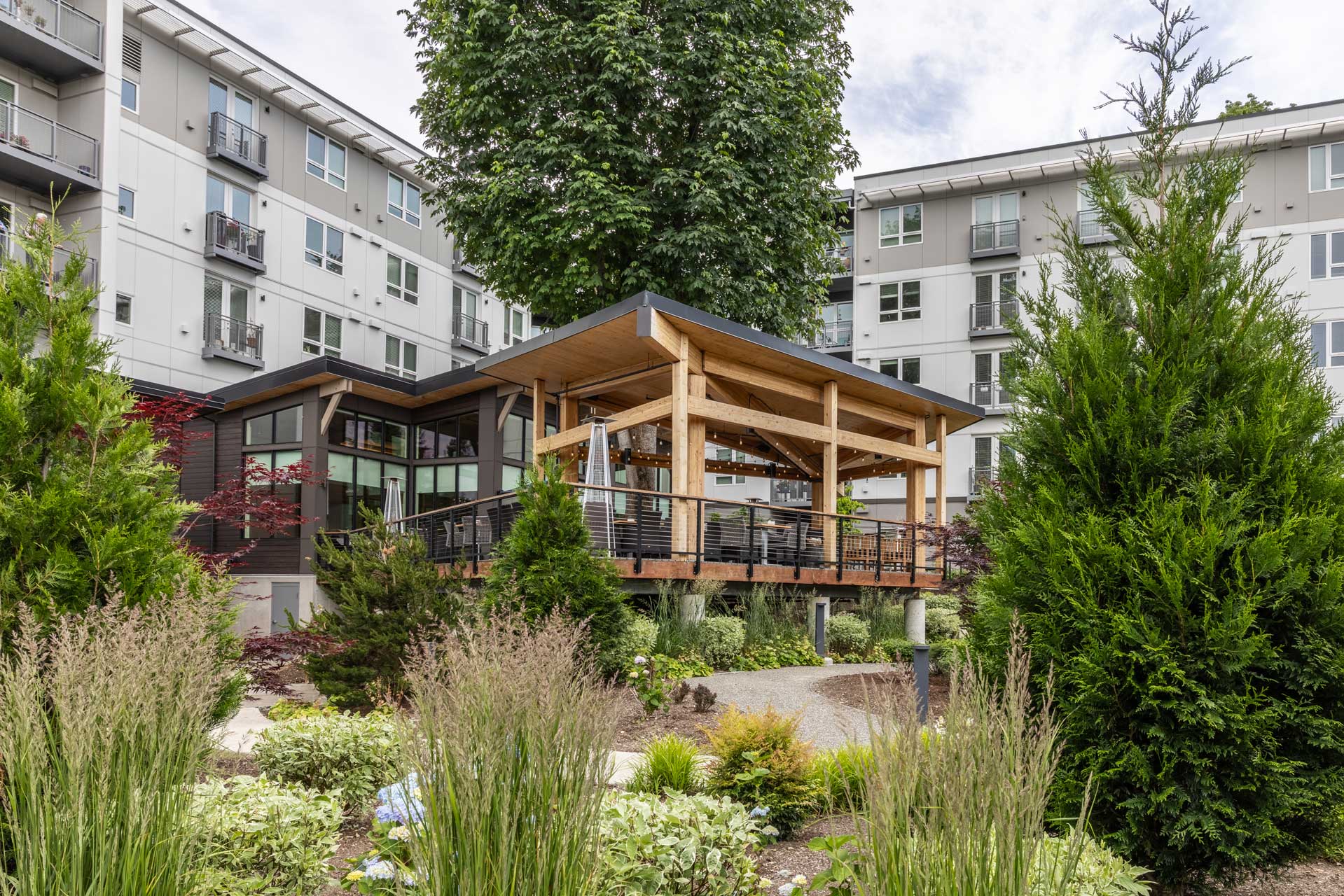 A modern wooden pavilion with a slanted roof and railings sits elevated among lush greenery at MorningStar Senior Living Kirkland, surrounded by contemporary apartment buildings—perfect for stylish retirement living.