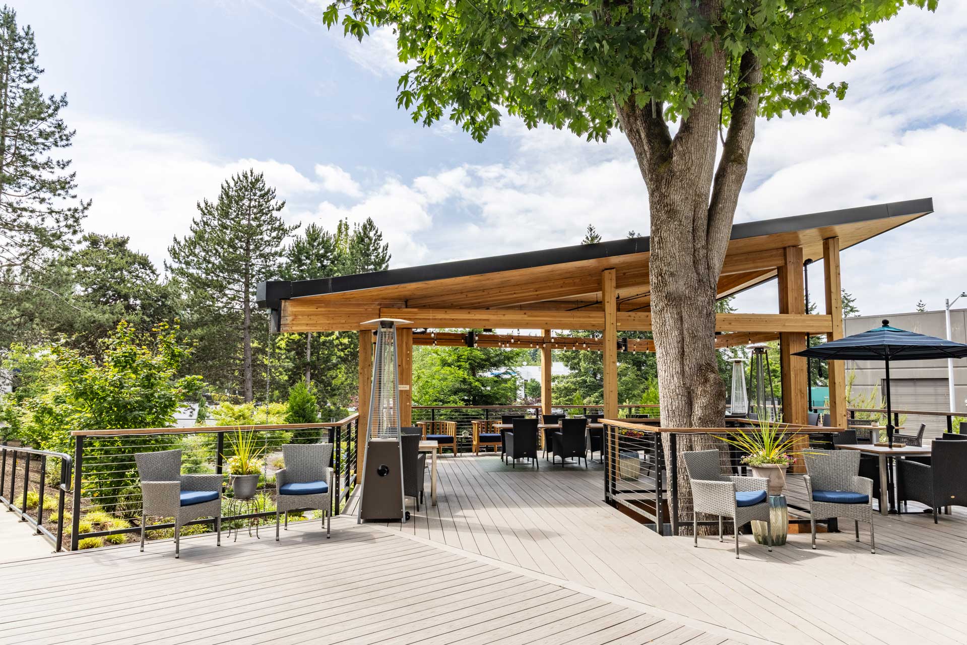 Modern outdoor patio with wooden decking and a canopy at MorningStar Senior Living Kirkland, featuring tables, chairs, potted plants, and large trees providing shade. The area offers tranquil Retirement Living surrounded by greenery under a partly cloudy sky.
