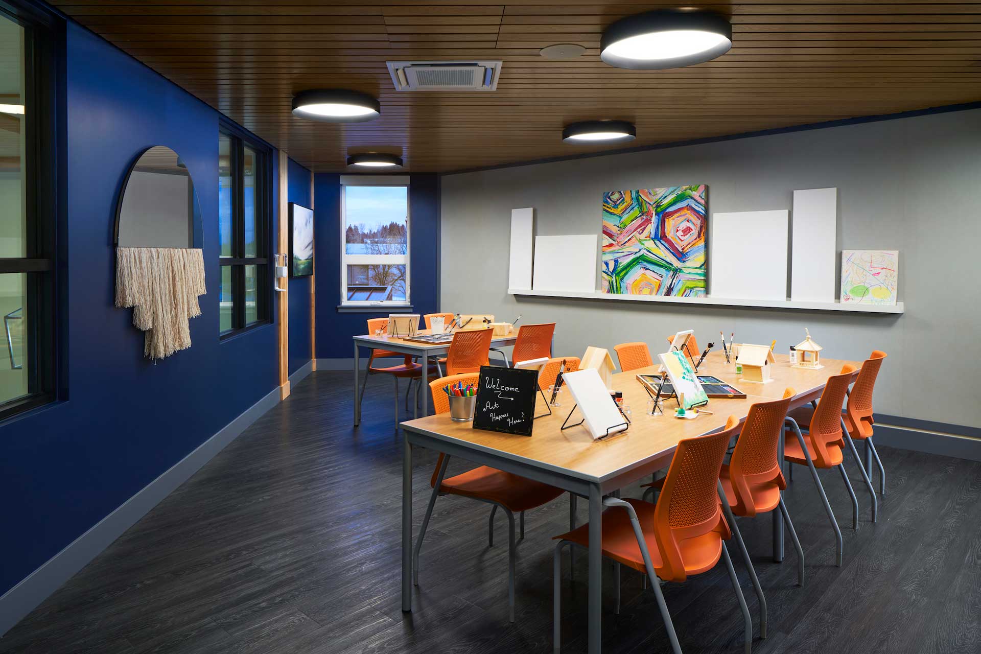 A modern art classroom at MorningStar Senior Living Kirkland features blue walls, orange chairs, wooden tables, and colorful paintings. Art supplies and easels are set up on the tables in this vibrant space for creative retirement living.