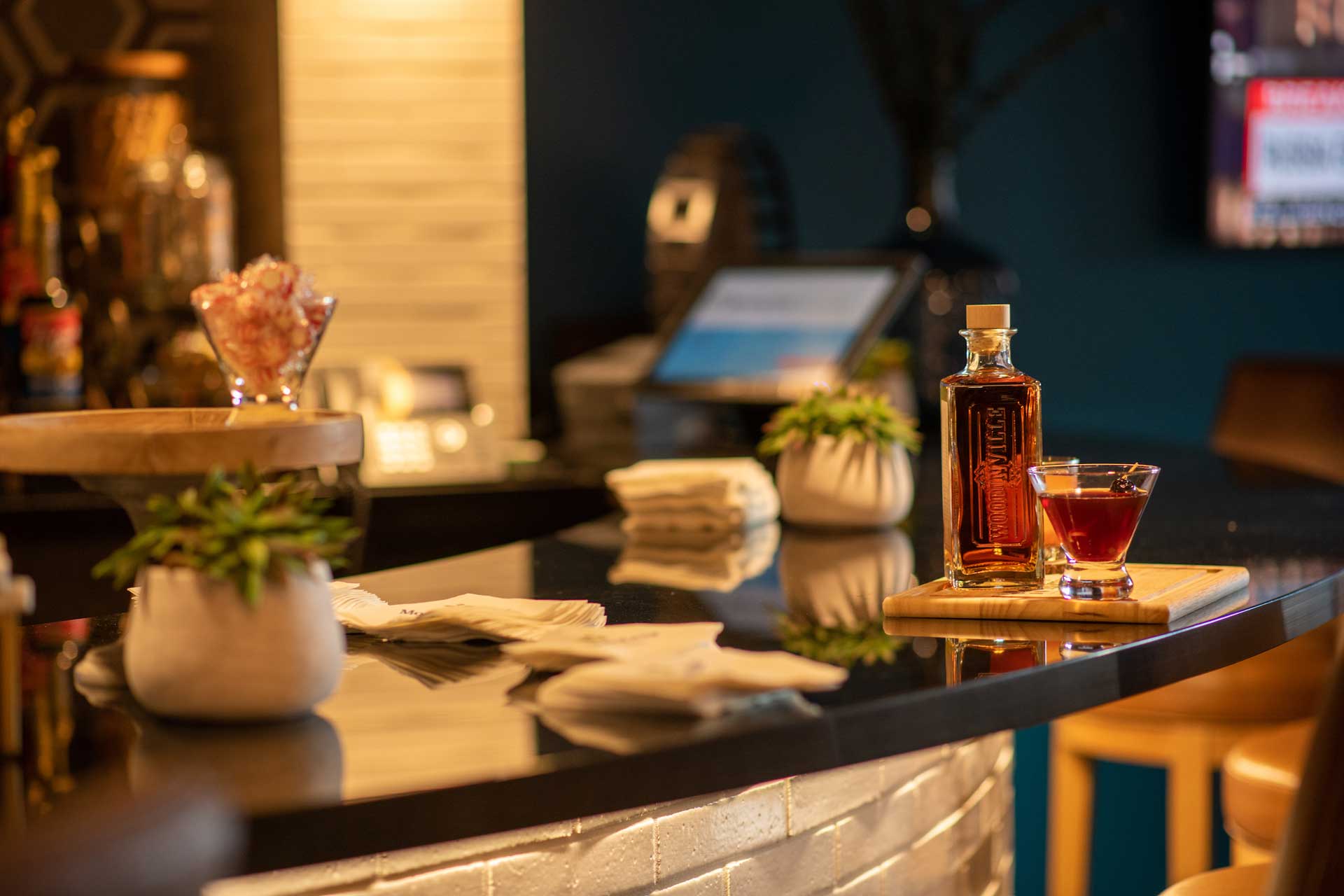 A bottle of whiskey and a glass cocktail sit on a wooden board atop a shiny bar counter at MorningStar Senior Living Kirkland, with potted plants, napkins, and bar equipment in a warmly lit, modern retirement living setting.