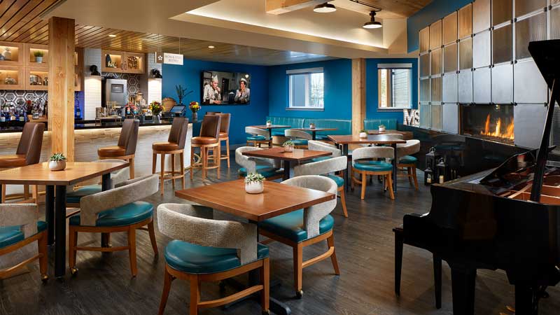 Modern restaurant interior at MorningStar Senior Living Kirkland features wooden tables, blue cushioned chairs, a bar area, TV, cozy fireplace, and black grand piano. Warm lighting enhances the inviting Retirement Living atmosphere.