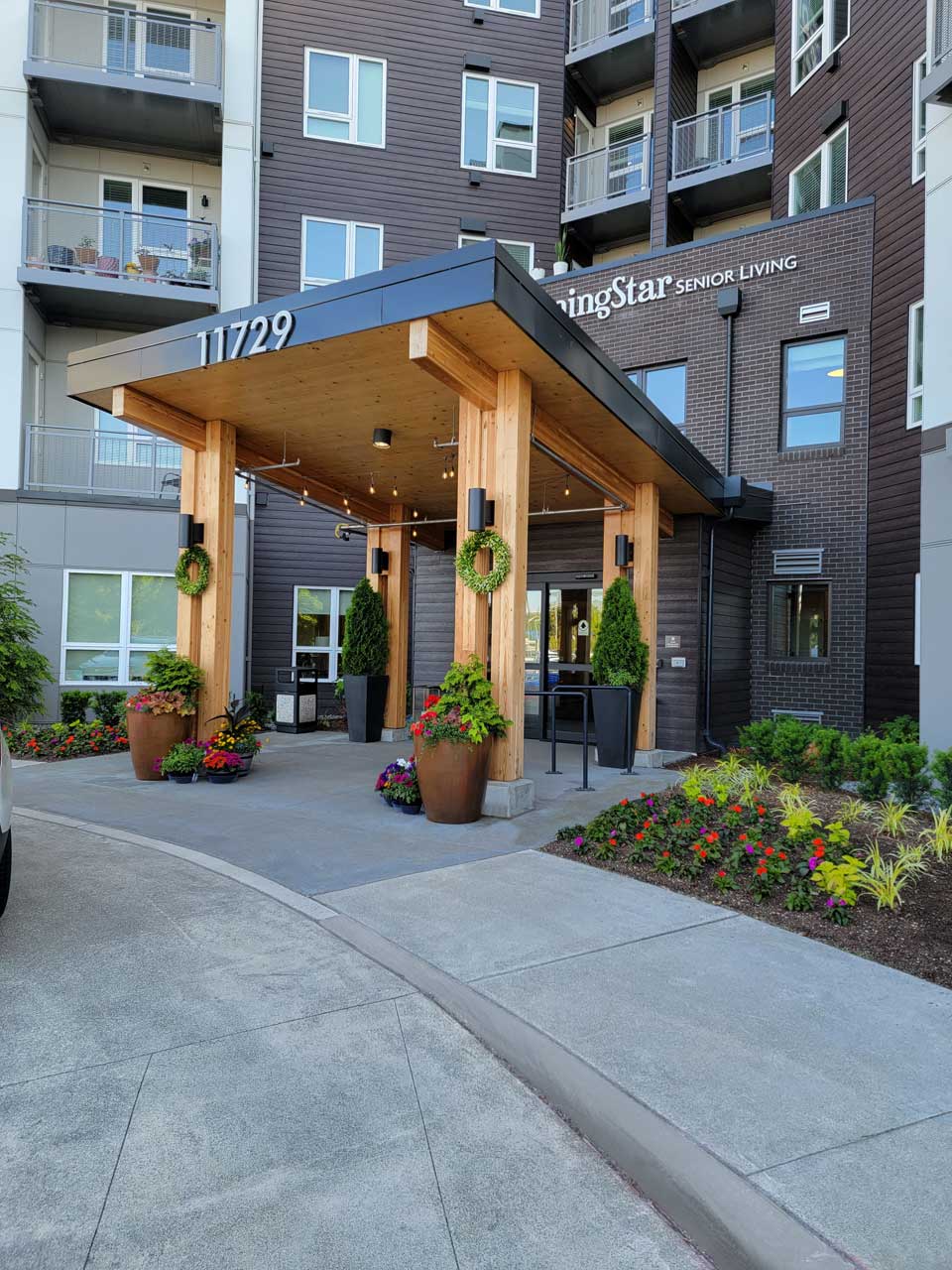 Entrance to MorningStar Senior Living Kirkland features a wooden awning, potted plants, green wreaths, and colorful flowers. This modern retirement living facility boasts large windows, balconies, and the address “11729” above the entrance.