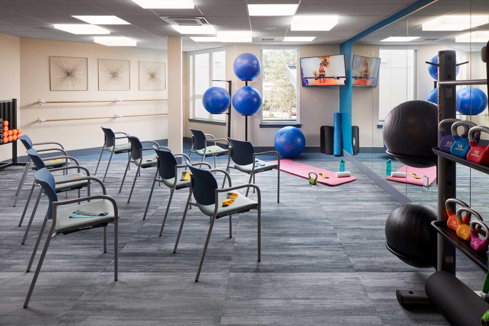 A bright fitness studio at MorningStar Senior Living Kirkland features chairs in rows, exercise balls, dumbbells, resistance bands, yoga mats, and a mirrored wall. Two TVs display workout videos as natural light fills this inviting retirement living space.
