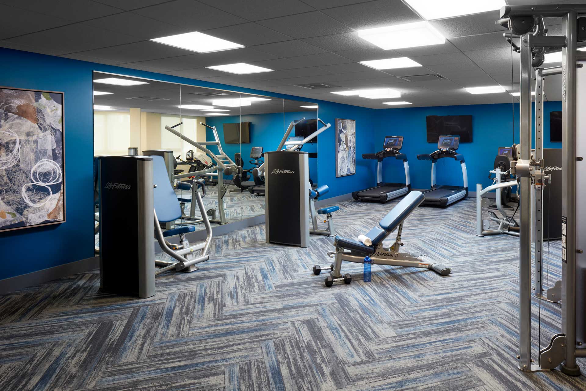 A modern gym at MorningStar Senior Living Kirkland features blue walls, chevron-patterned carpet, treadmills, weight machines, benches, and a large mirrored wall—creating an inviting space for active retirement living. Abstract art and bright lights complete the room.