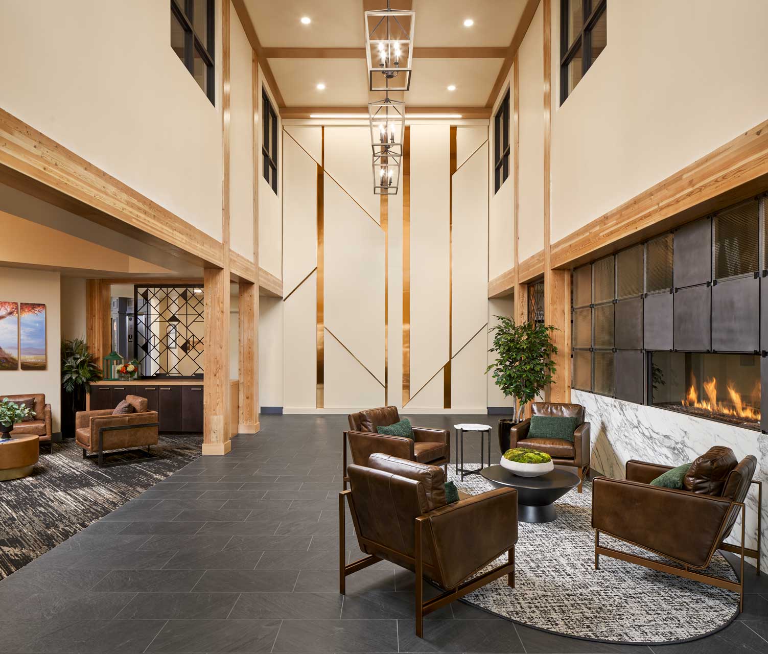 Modern hotel lobby with high ceilings, leather armchairs around a circular table, a large fireplace, indoor plants, and contemporary artwork on the walls evokes the inviting atmosphere found at MorningStar Senior Living Kirkland Retirement Living.