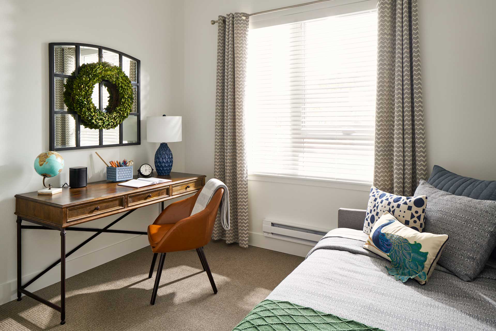 A cozy bedroom at MorningStar Senior Living Kirkland features a wooden desk, brown chair, globe, lamp, and clock by a window with gray curtains—perfect for comfortable retirement living. A wreath hangs above the desk near the inviting bed.
