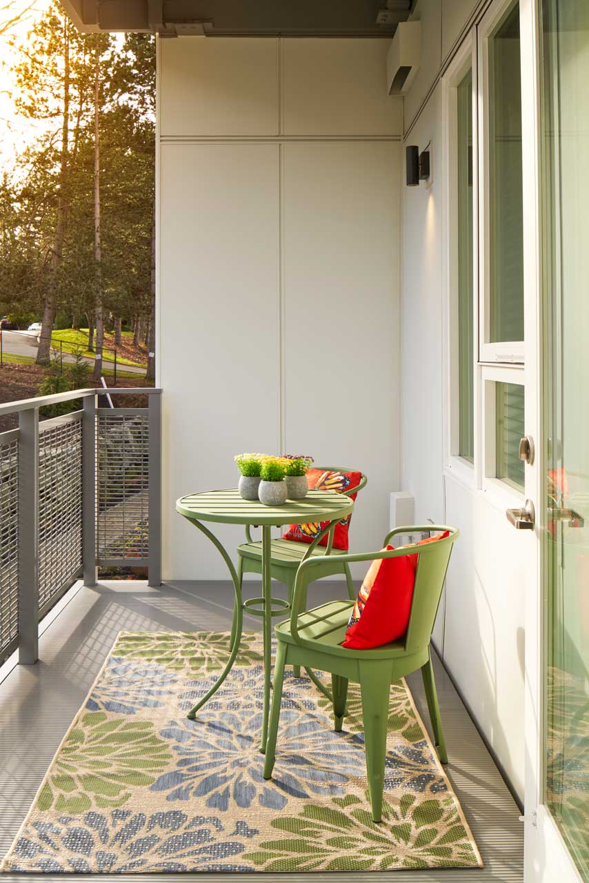 A small balcony at MorningStar Senior Living Kirkland features a green metal table, two green chairs with red cushions, a patterned rug, and potted plants. Perfect for enjoying Retirement Living while overlooking trees in the warm evening light.