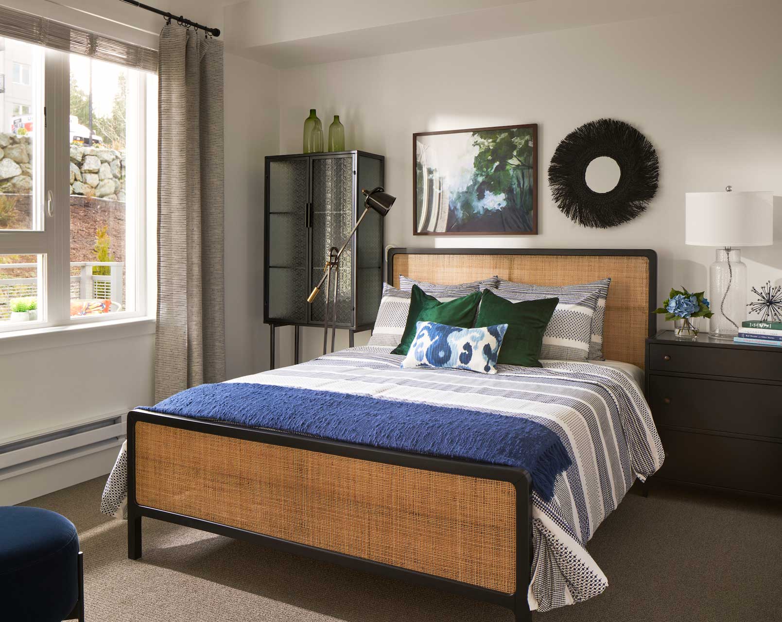 A modern bedroom at MorningStar Senior Living Kirkland features a woven headboard, blue and gray bedding, striped pillows, a black nightstand with a white lamp, artwork above the bed, a tall glass cabinet, and a window with gray curtains—perfect for retirement living.