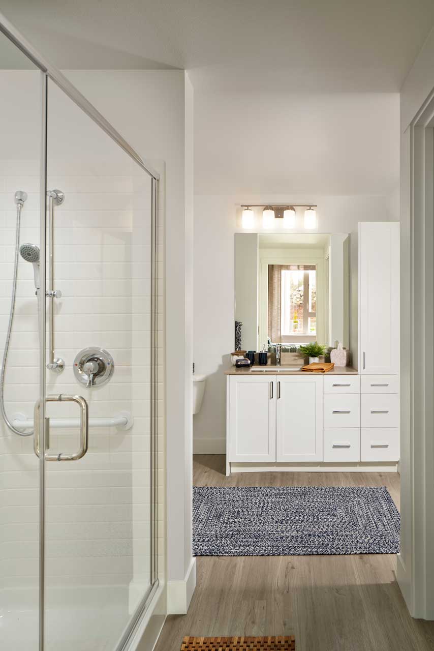 A modern bathroom at MorningStar Senior Living Kirkland features a glass shower on the left, a white vanity with storage, a large mirror and sink, plus a window above. The light wood-style flooring and blue patterned rug enhance retirement living comfort.