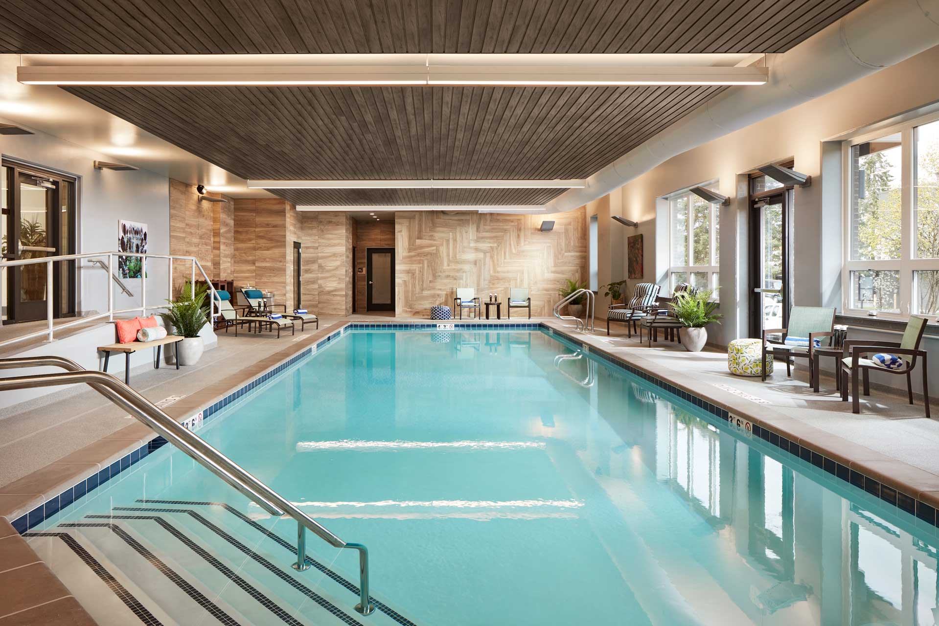 Enjoy the indoor swimming pool at MorningStar Senior Living Kirkland, where clear water, lounge chairs, and potted plants create a relaxing retirement living atmosphere filled with natural light and warm wooden accents.