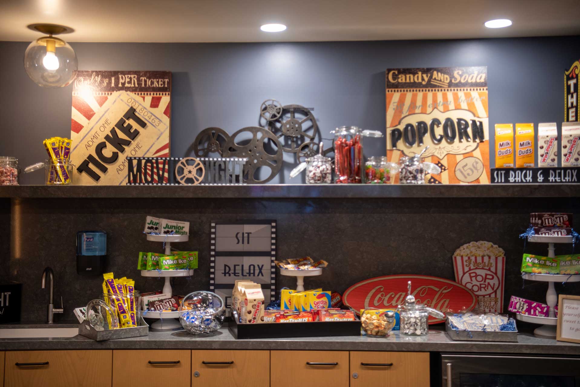 A home theater snack bar at MorningStar Senior Living Kirkland features shelves with popcorn, candy, a Coca-Cola sign, and movie-themed decor like film reels and marquee signs—offering a fun treat for residents enjoying retirement living.