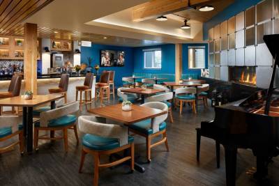 Modern restaurant interior at MorningStar Senior Living Kirkland, featuring wooden tables and chairs, teal accents, a bar area, grand piano, wall art, and a blue accent wall with fireplace—perfect for vibrant retirement living.