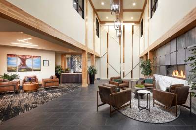 A modern lobby at MorningStar Senior Living Kirkland features high ceilings, wood beams, and large windows. Leather chairs surround a table near the fireplace, with plants and artwork creating a well-lit, inviting space perfect for retirement living.