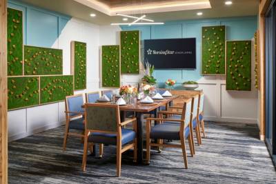 A modern dining room at MorningStar Senior Living Kirkland features a wooden table set for eight, blue cushioned chairs, floral centerpieces, green wall art, and a sign reading "MorningStar Senior Living" on the back wall—perfect for retirement living.