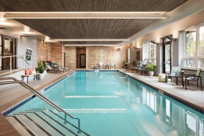 Indoor swimming pool with metal handrails, lounge chairs, potted plants, and large windows letting in natural light at MorningStar Senior Living Kirkland. The wooden ceiling and modern decor create a welcoming retirement living space.