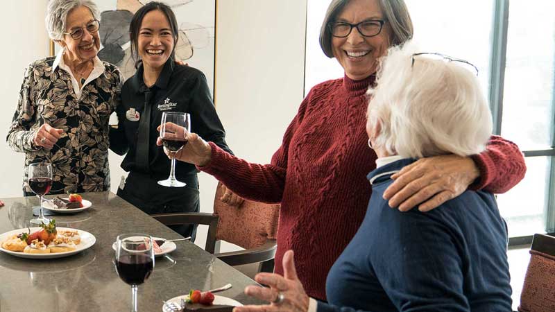 Four women, three older and one younger, stand and sit around a table, smiling and laughing together. At MorningStar Senior Living Kirkland, their shared meal and glasses of red wine create a warm, friendly Retirement Living atmosphere.