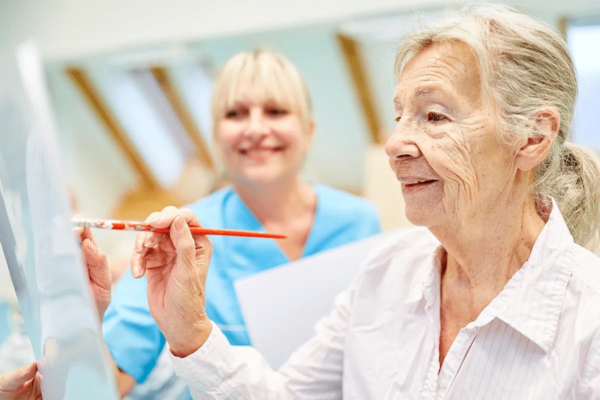 An elderly woman smiles while painting on a canvas, holding a paintbrush. A nurse in blue scrubs stands behind her, also smiling. The bright, cheerful setting reflects the vibrant community at MorningStar Senior Living Kirkland.