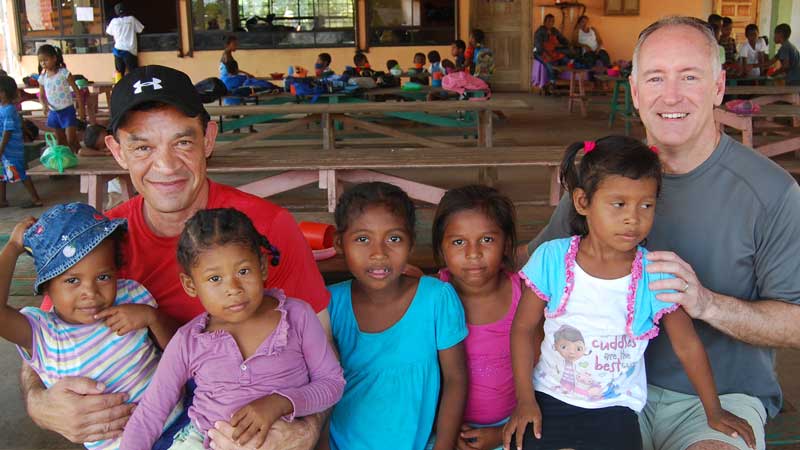 Two men from MorningStar Senior Living Kirkland sit with five young girls on their laps and beside them, all smiling. In the background, more children gather at tables in a spacious room filled with benches and colorful bags.