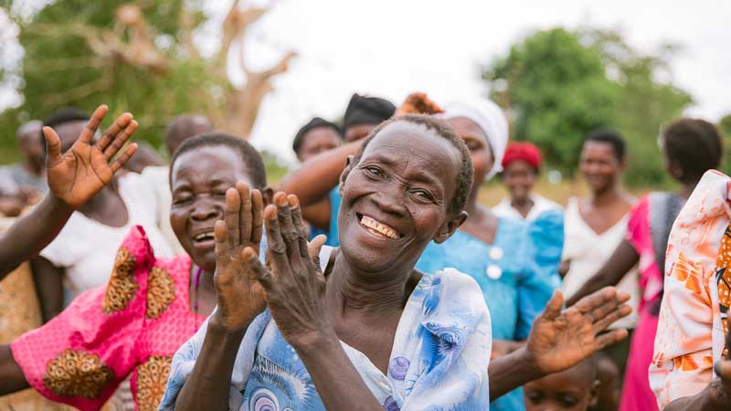 A group of people outdoors, smiling and clapping joyfully. The focus is on an older woman in a blue patterned dress, surrounded by others in colorful clothing—capturing the vibrant spirit of Retirement Living at MorningStar Senior Living Kirkland.