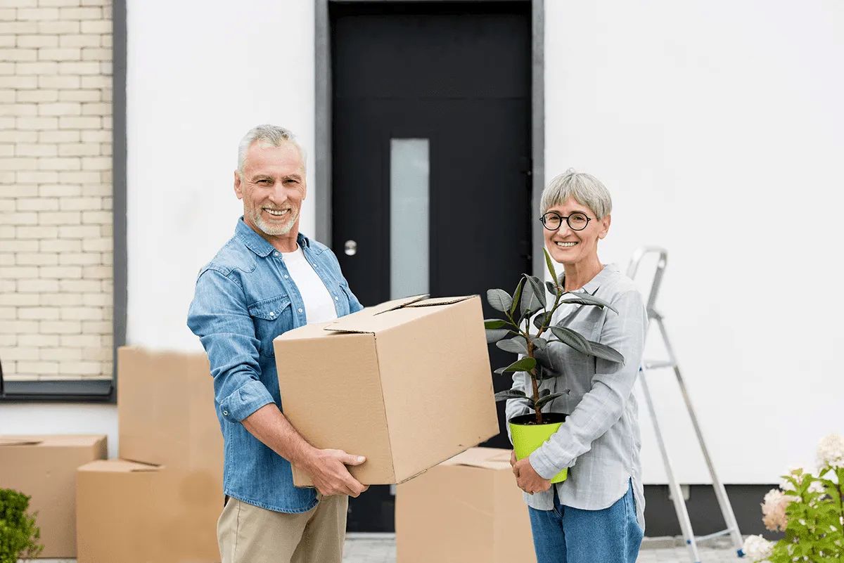 Smiling older couple stands outside a house, holding a cardboard box and a potted plant. Surrounded by moving boxes, they look ready to start their next chapter—perhaps with Retirement Living at MorningStar Senior Living Kirkland.