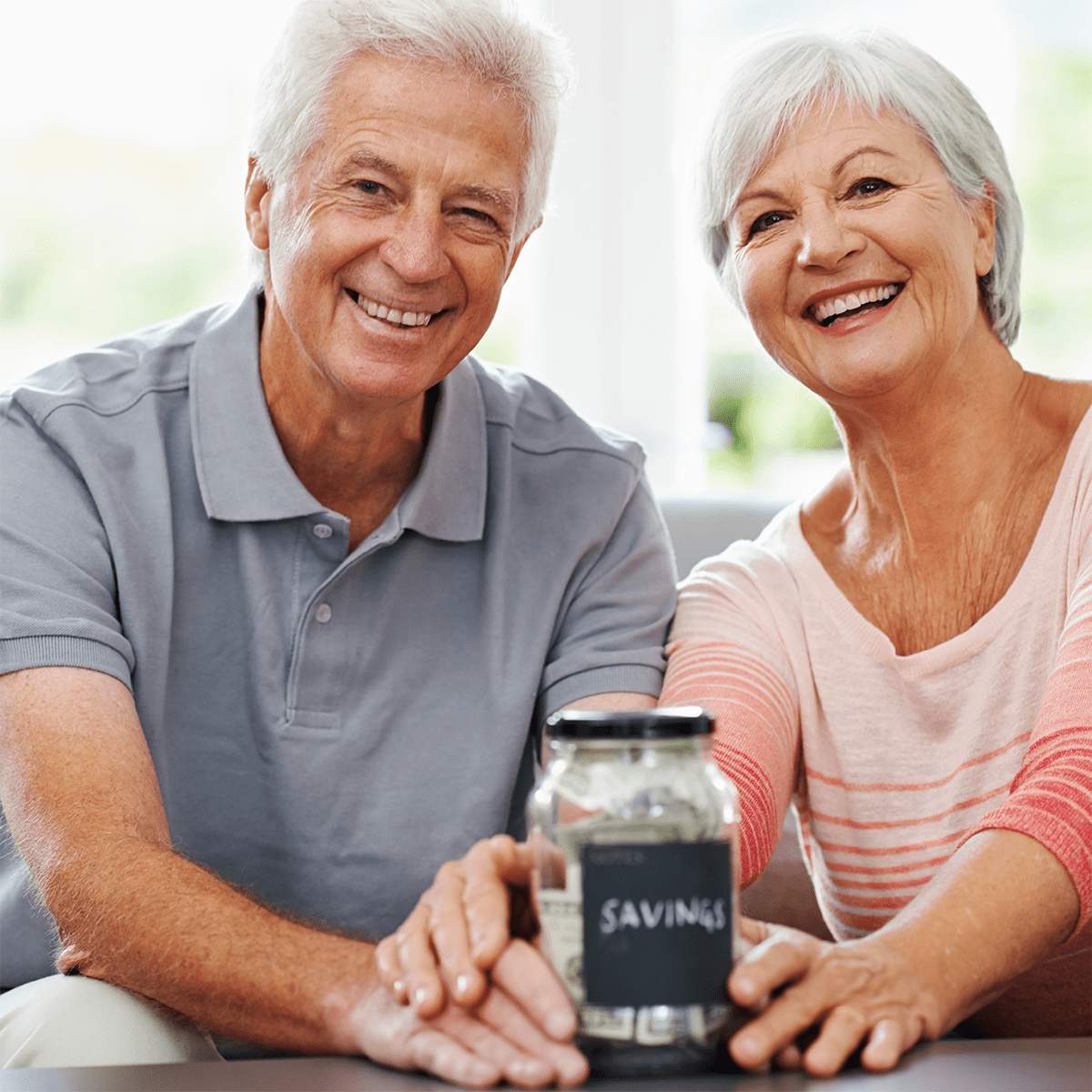 Senior-Savings Smiling older couple sitting together, holding a glass jar labeled "SAVINGS" filled with cash, suggesting financial planning or saving for the future at MorningStar Senior Living Kirkland.