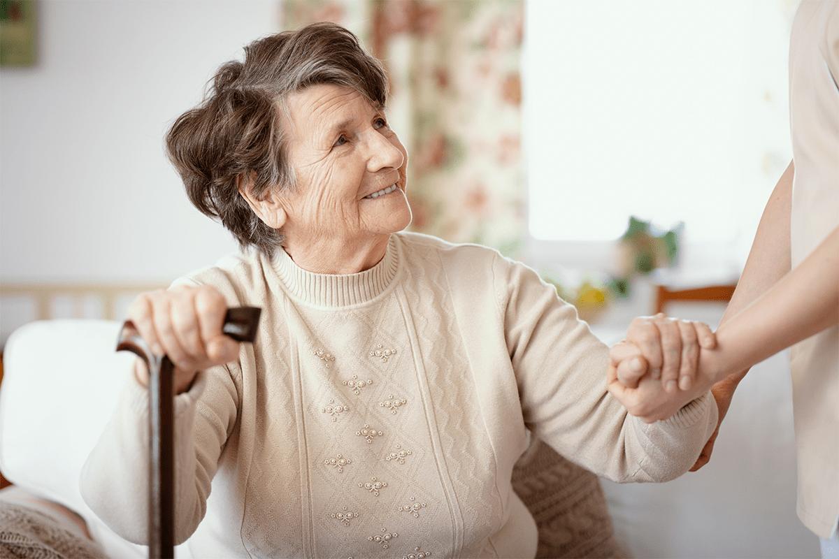 An elderly woman with short gray hair smiles while holding a cane and the hand of another, sitting indoors with floral curtains—a warm moment of connection at MorningStar Senior Living Kirkland.