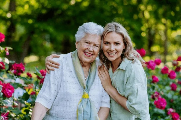 Two women, one elderly and one younger, smile and embrace in a colorful garden filled with blooming flowers at MorningStar Senior Living Kirkland. The lush, green background highlights the joy of retirement living on a sunny, pleasant day.
